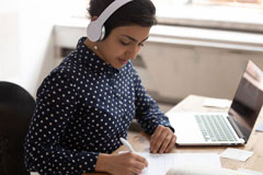 Indian ethnicity woman wearing headphones listens educational course writing down necessary useful information gain knowledge online use internet lesson, interpreter do translation sit at desk indoors